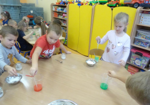 Troje dzieci nabiera pipetkami zabarwioną na pomarańczowo oraz na zielono ciecz, którą przenoszą do słoiczków z roztworem z soli.